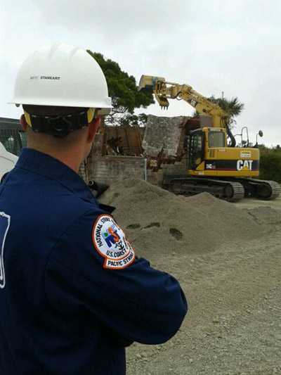 Taken by Petty Officer 3rd Class Blaine Meserve-Nibley. Chief Petty Officer Cody Staneart, a response technician with the U.S. Coast Guard's Pacific Strike Team, supervises the demolition of a house on Indian Island in Eureka, CA. The house was recently abated of asbestos and human remains were discovered under the old foundation. The Coast Guard was on scene to fill the Environmental Protection Agency's request for trained personnel to conduct on-scene safety, particularly pertaining to waterside operations.